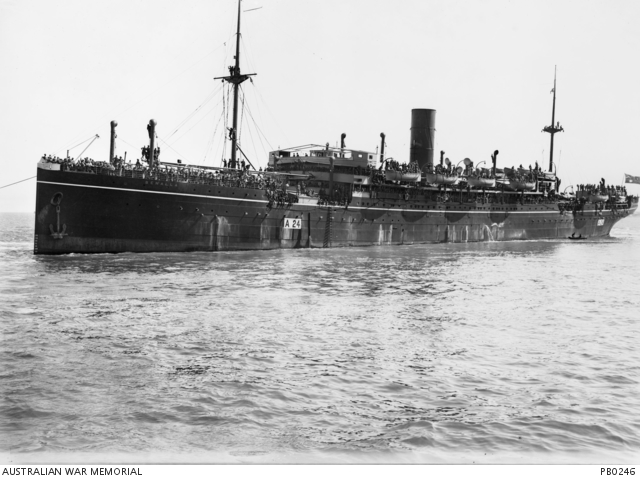 A large, dark coloured ship with a single funnel crowded with troops, departing from the wharf. A small rowing boat is near the stern of the ship.